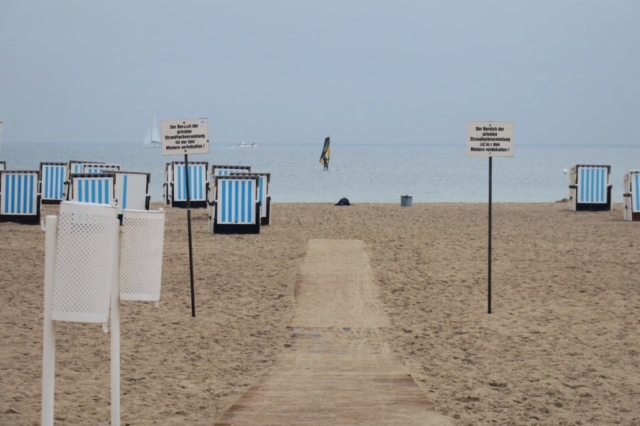 Strand, leere Strandkörbe, ein Surfer auf ruhigem Wasser.