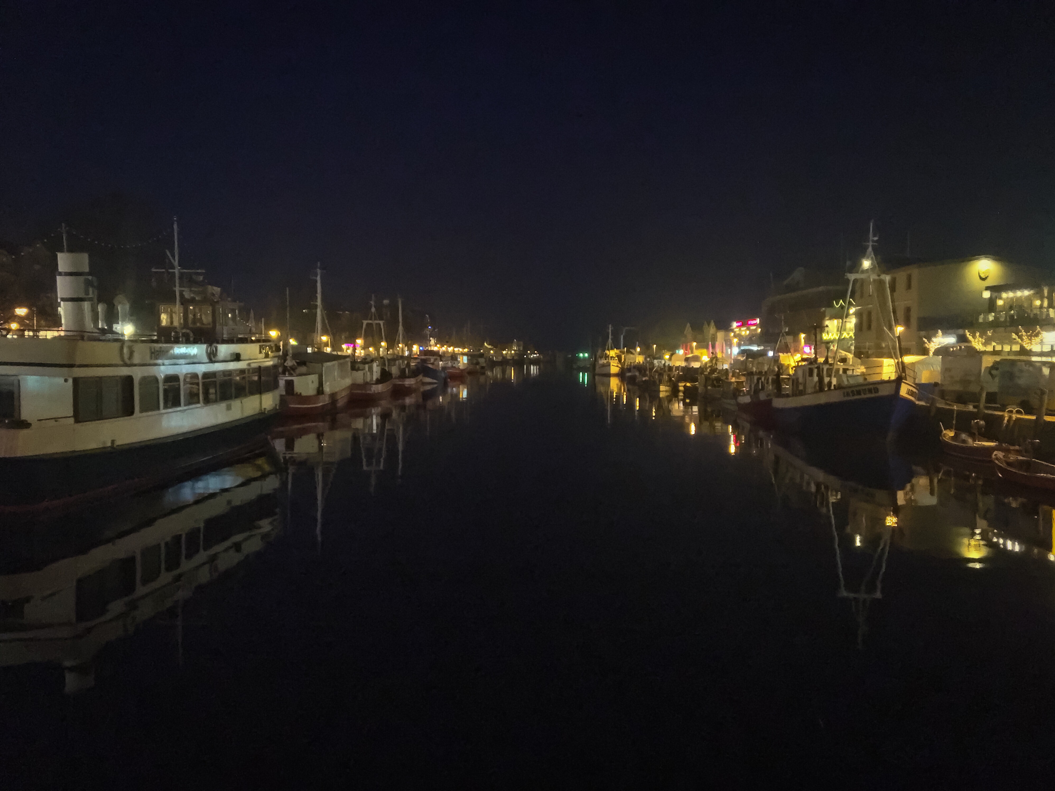 Alter Strom Warnemünde bei Dunkelheit. Schiffe und Leuchtreklamen spiegeln sich im glatten Wasser.