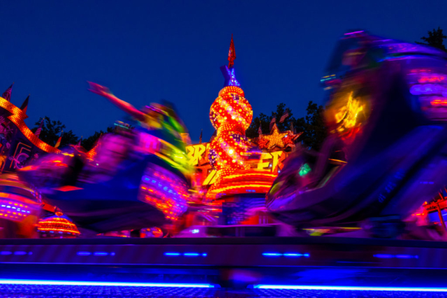 Langzeitbelichtung: Karussell-Gondeln ziehen bunte Leuchtspuren durch die Abenddämmerung.