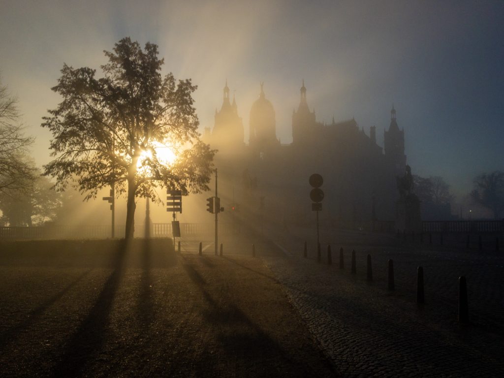 Morgensonne leuchtet durch Nebel und Dunst am Schweriner Schloss. Es bilden sich goldene "Strahlen" im Dunst.