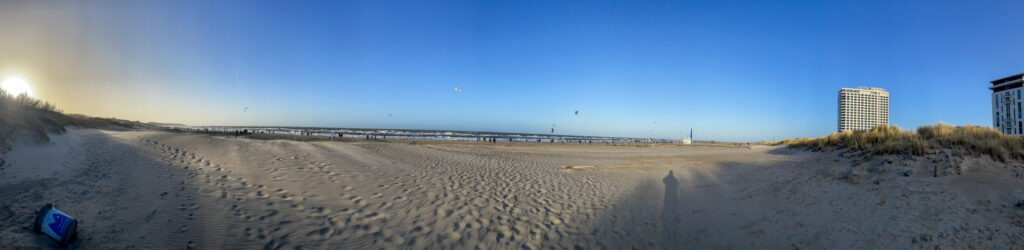 Panoramaaufnahme eines Strandaufgangs von Warnemünde, links geht die Sonne hinter Dünen unter, auf dem Wasser sind viele bunte Kitesurfer-Drachen zu sehen, rechts steht das Hotel Neptun im Sonnenlicht.