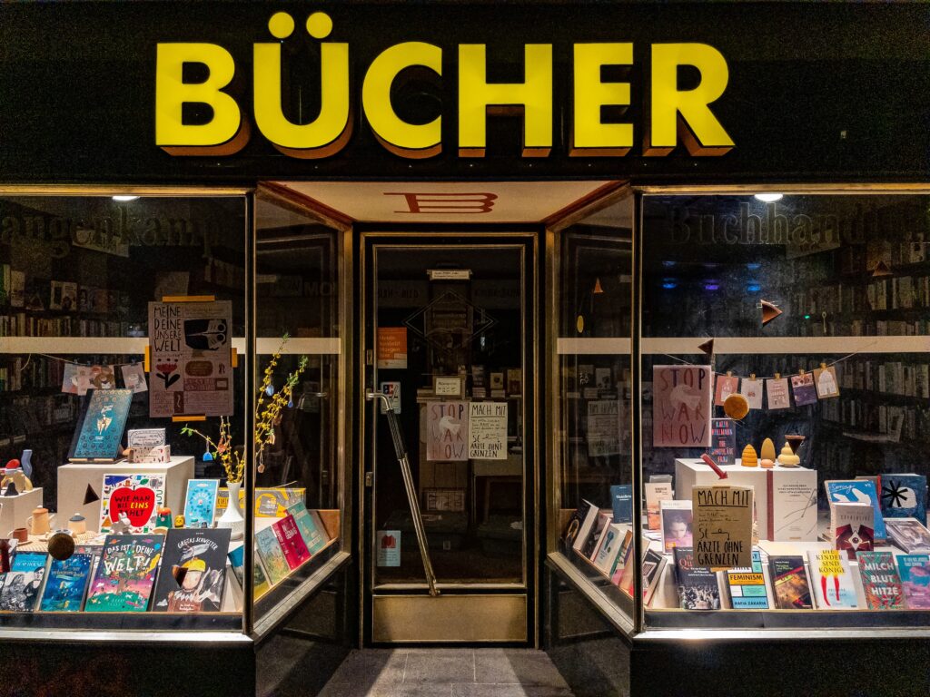 Nachtaufnahme. Hell erleuchtete Schaufenster und mittig die Eingangstür einer Buchhandlung. Bunte Auslagen in den hoch aufragenden Schaufenstern. Über dem Eingang prangt in serifenlosen Leuchtbuchstaben das Wort „Bücher“.