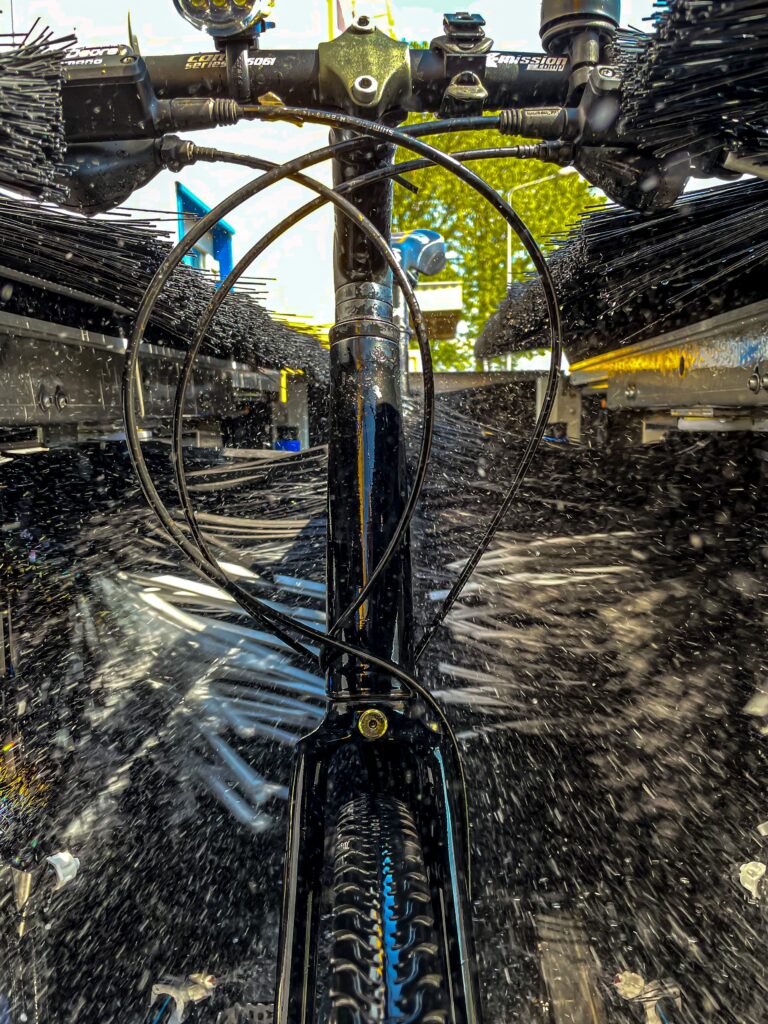 Blick in eine Fahrradwaschanlage: Fahrrad von vorn. Schwarze Bürsten rotieren, tausende Wassertropfen spritzen, der schwarz lackierte Lenker glänzt nass.