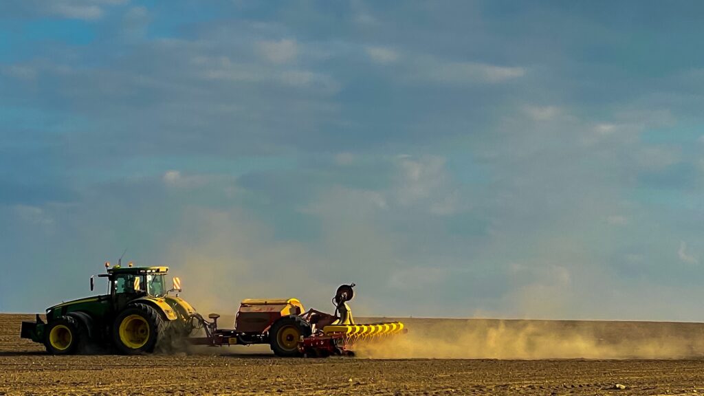 Ein grün-gelber Traktor zieht eine Landmaschine über ein sandiges Feld, auf dem keine Pflanzen stehen. Vor dem locke bewölkten blauen Himmel zeichnet sich eine lange grau-braune Staubwolke ab, die das Gefährt aufgewirbelt hat.