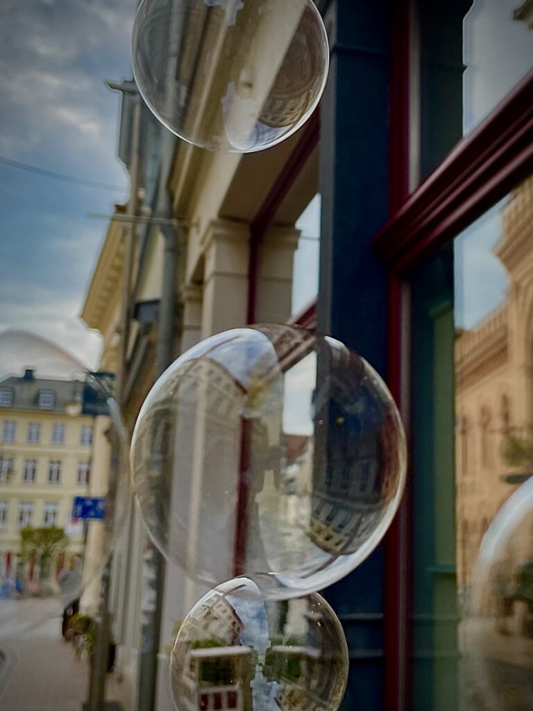 Seifenblasen schweben vor einem großen Fenster, in der durchsichtigen Haut sind rund verzerrte Reflexionen von Hausfassaden zu erkennen.