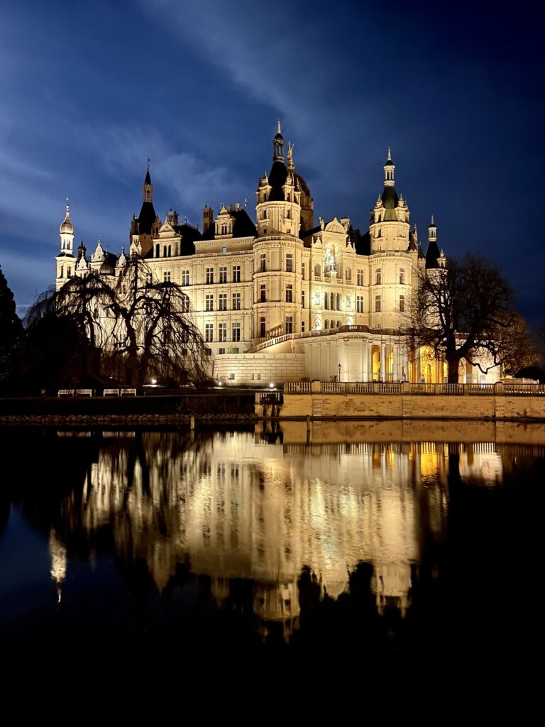 Die angeleuchtete ockerfarbene Fassade des Schweriner Schlosses spiegelt sich nachts auf der fast glatten Wasseroberfläche des Sees. Die Wolken am dunkelblauen Nachthimmel werden sanft vom Mondlicht erhellt.