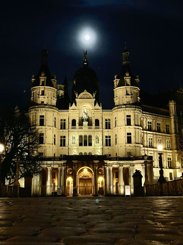 Blick über die Schlossbrücke zum Schweriner Schloss bei Nacht. Darüber leuchtet weiß der fast volle Mond.